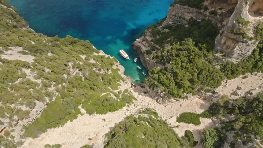 A stunning aerial flyover reveals a secluded turquoise bay where boats are anchored at the end of a dramatic, rocky gorge