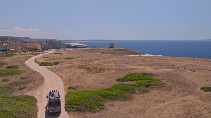 An aerial view shows a camper van driving on a scenic dirt road along an arid coastline towards the blue ocean on a sunny day