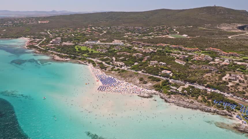 An expansive aerial view captures a bustling beach with colorful umbrellas and swimmers in the vibrant turquoise waters of a Mediterranean coastline