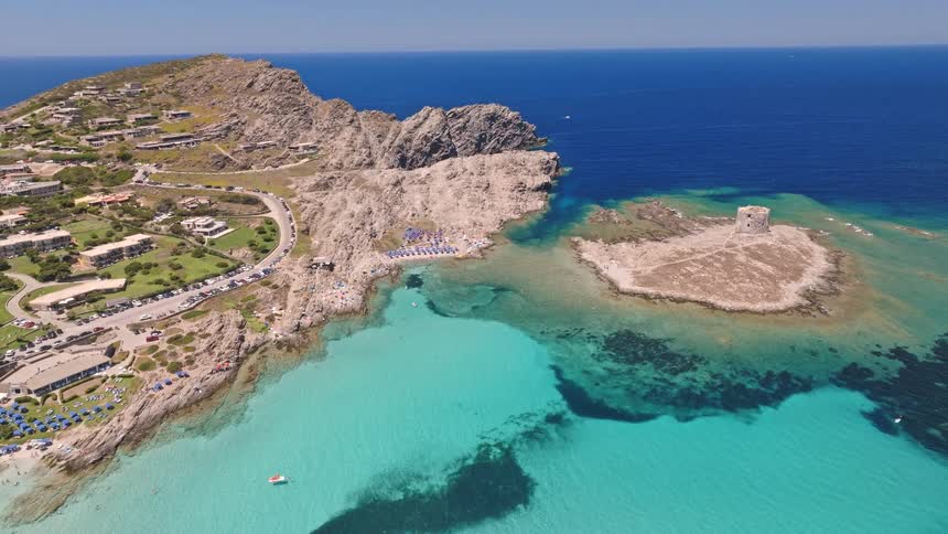 A stunning aerial pull-back shot reveals the iconic La Pelosa beach in Sardinia, with its ancient stone tower, rocky coastline, and vibrant turquoise sea