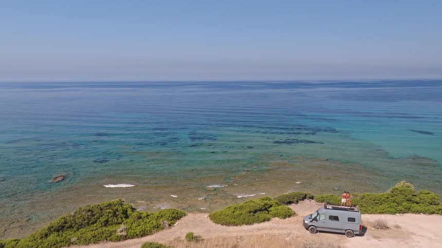 A stunning aerial view pulls back to reveal a couple on their camper van, parked on a cliff and admiring the vast turquoise ocean