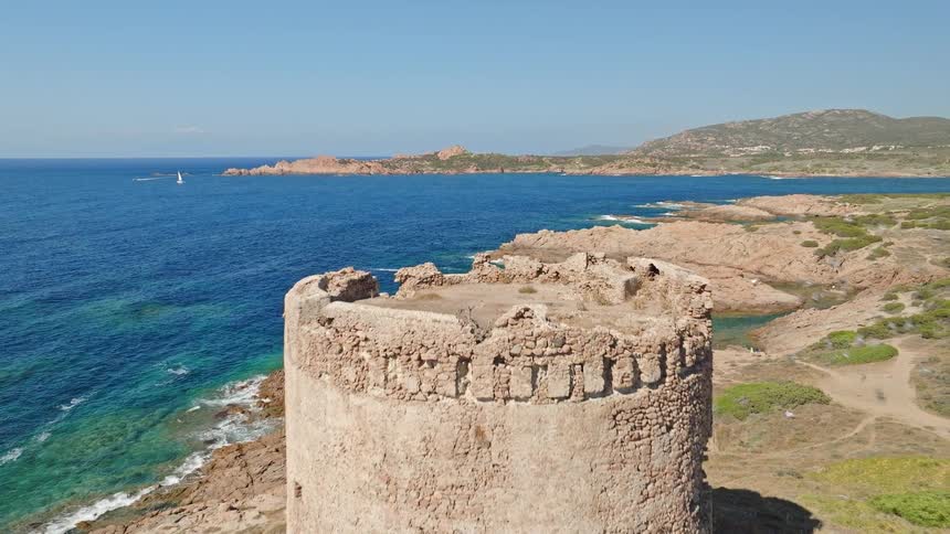 Aerial pull-back shot reveals an ancient stone watchtower overlooking the rocky coastline and turquoise sea on a sunny day