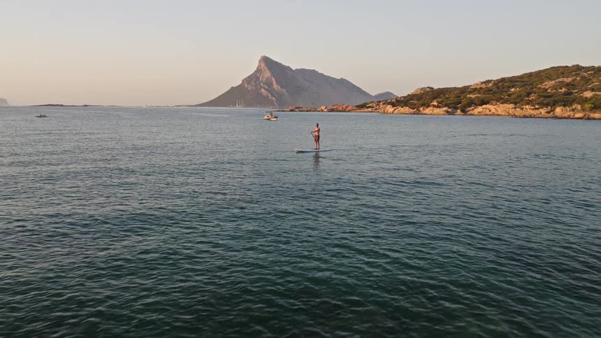 Drone shot reveals a man stand-up paddleboarding near a beautiful rocky shoreline with a mountain in the background at sunset