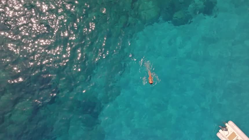 An aerial top-down drone shot shows a woman floating and swimming in calm, crystal-clear turquoise ocean water next to small boats