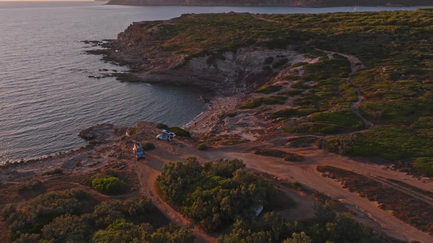 A dramatic aerial shot pulls back to reveal two camper vans parked on a rugged coastal cliff as the sun sets over the ocean