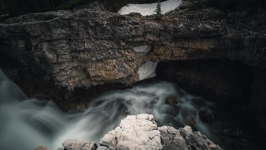 A breathtaking long exposure timelapse reveals a powerful waterfall carving its way through a rocky, snow-covered mountain landscape
