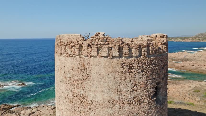 Drone ascends over an ancient stone watchtower on a rocky coastline, revealing the stunning turquoise sea as a flock of birds takes flight