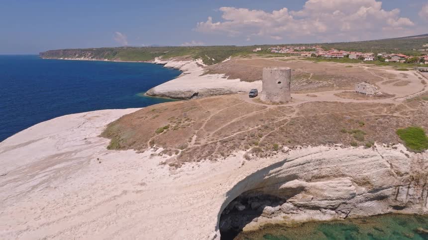 Breathtaking aerial pull-back shot of a camper van parked beside a historic stone tower on a dramatic white cliff coastline overlooking the turquoise sea