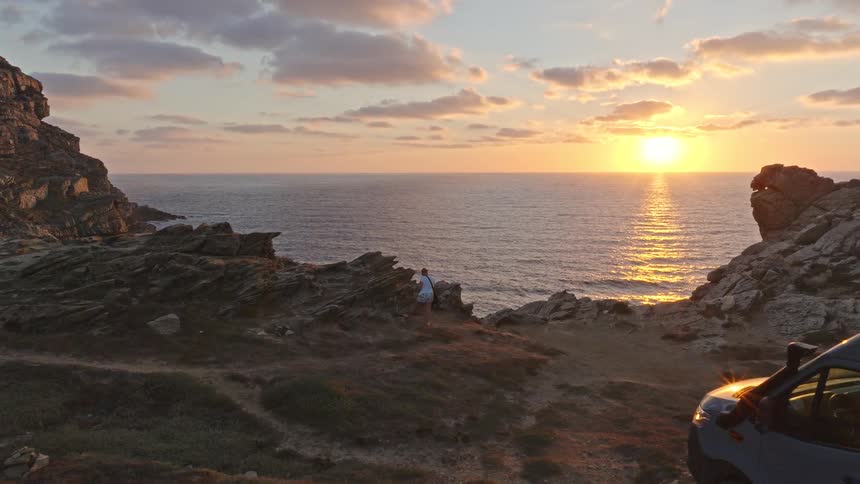 A drone flies past a camper van parked on a rugged cliffside as people watch the golden sunset over the vast ocean