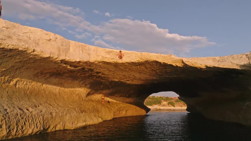 A young man performs a thrilling slow-motion jump from a natural rock arch into the sea during a beautiful golden hour
