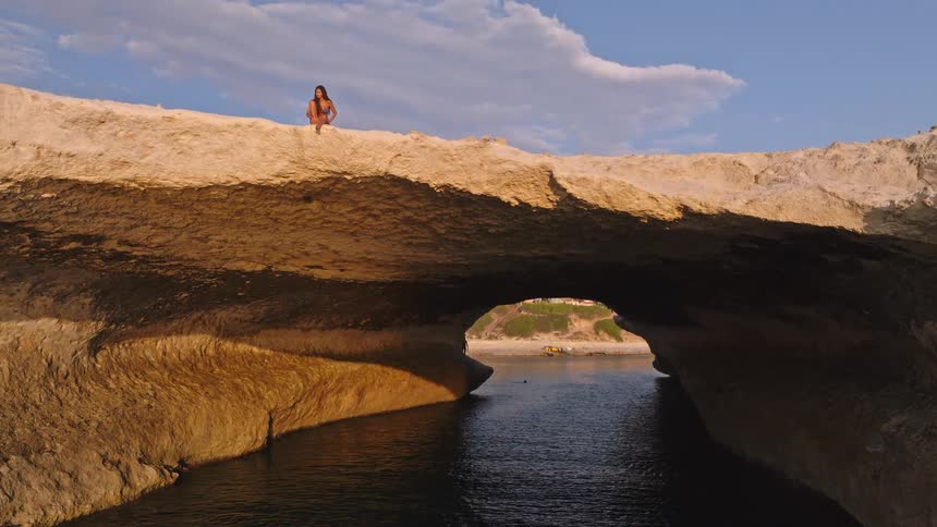 A woman meditates peacefully on top of a magnificent natural rock arch over the ocean at golden hour