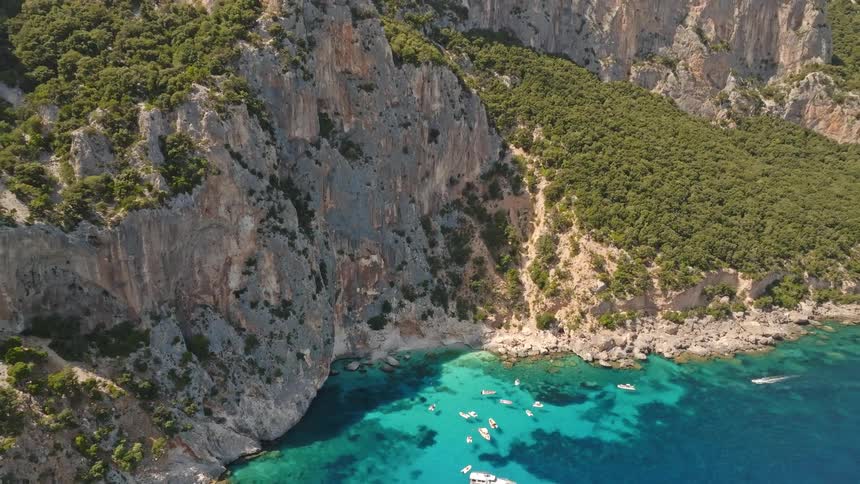 A stunning aerial view reveals a hidden cove with crystal clear turquoise water, anchored boats, and towering green cliffs