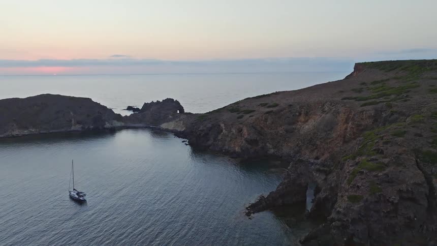 An aerial view reveals a lone sailboat peacefully anchored in a secluded, rocky cove as the sun sets over the ocean