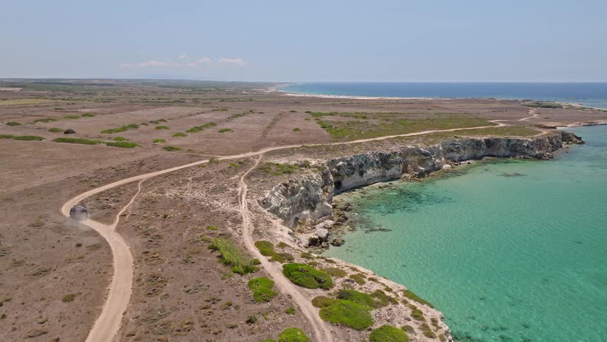 A drone follows an SUV as it kicks up dust on a scenic dirt road along a rugged cliffside overlooking the turquoise Mediterranean Sea