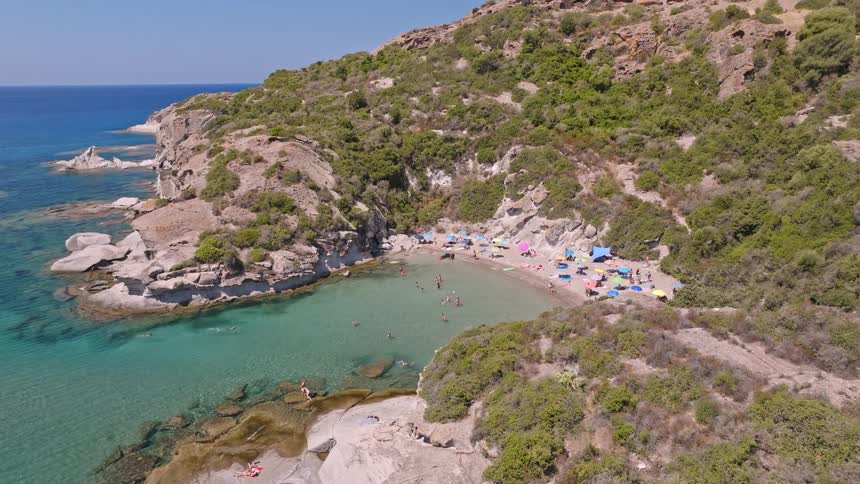 An aerial drone shot slowly pushes in on a scenic hidden cove where people swim in turquoise water and relax on a sandy beach