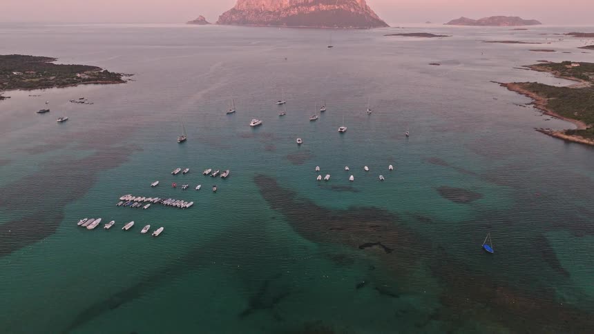 A stunning aerial view flies over a tranquil turquoise bay filled with boats, revealing the magnificent Tavolara Island at sunset