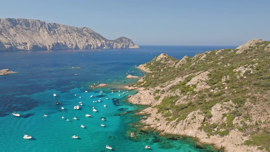 An idyllic aerial shot orbiting a verdant island surrounded by clear turquoise water and numerous yachts on a sunny day
