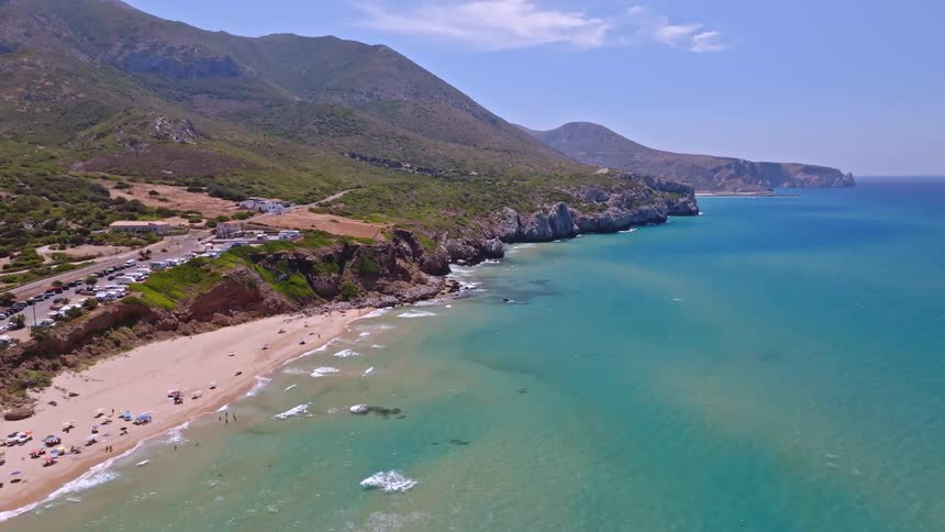 Aerial shot of a beautiful sandy beach with turquoise water, backed by a mountainous coastline and enjoyed by tourists on a sunny day