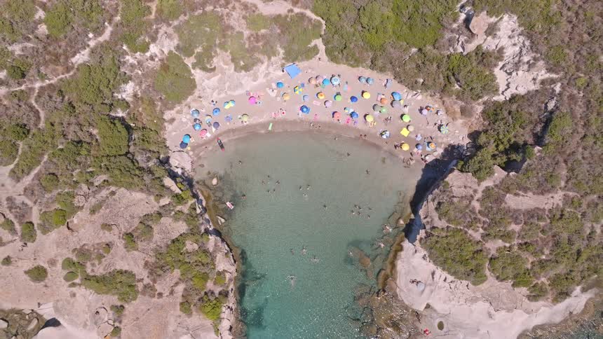A top-down aerial view slowly pulls back from a small, secluded sandy cove crowded with colorful beach umbrellas and people enjoying the clear blue water