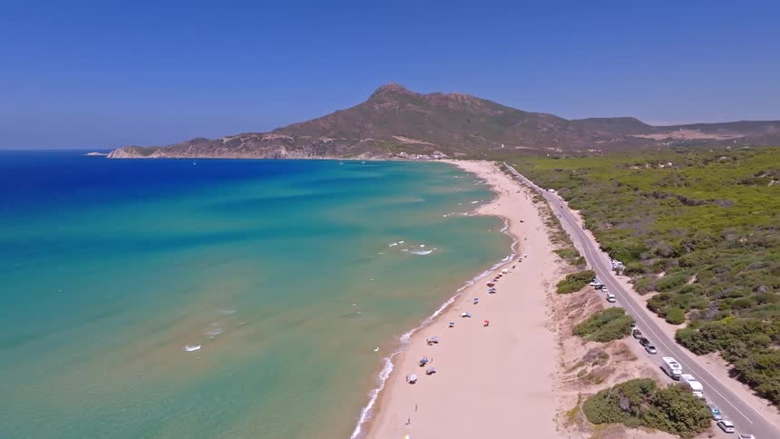 Aerial drone footage shows a stunning turquoise ocean meeting a long sandy beach next to a coastal highway with a majestic mountain in the background
