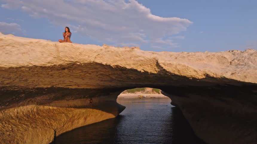 Slow aerial pullback from inside a sea cave reveals a woman meditating atop a magnificent white rock arch over the ocean at golden hour