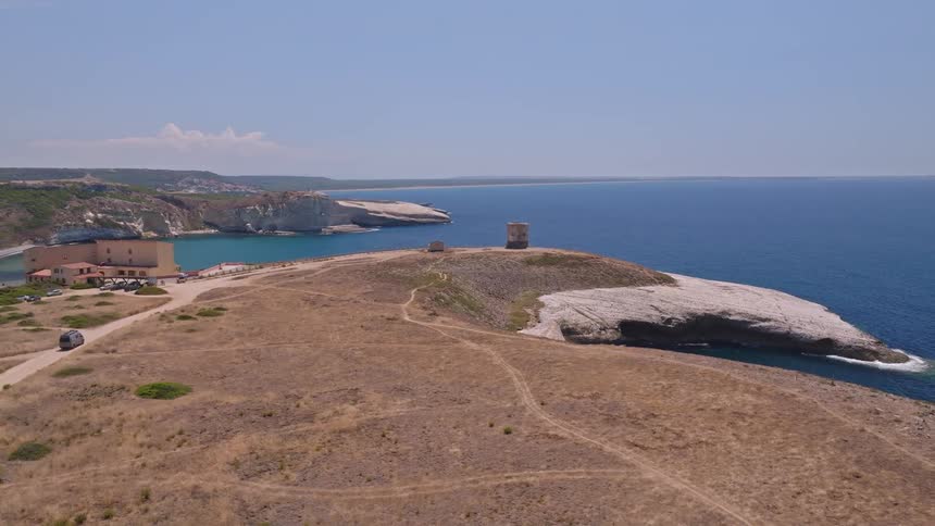 A drone follows a camper van driving along a rugged coastal road toward a historic stone tower perched on a white cliff headland overlooking the sea