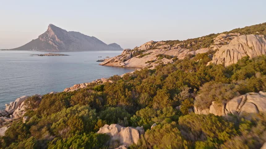 Drone flies along a picturesque rocky coastline with turquoise coves and a distant mountain island during a warm sunset