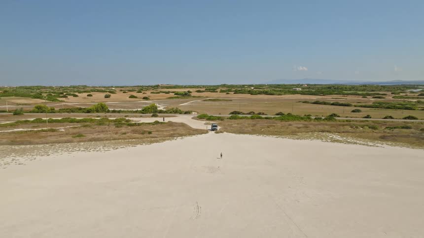 A lone woman jogs across a vast sandy expanse toward her camper van as the drone pulls back to reveal the epic natural landscape