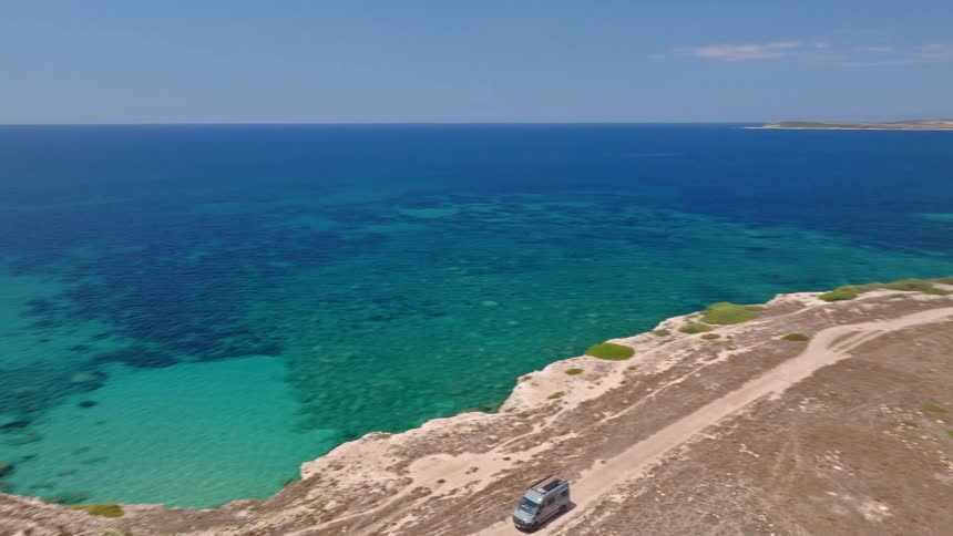Drone follows a camper van driving along a rugged coastal dirt road with stunning views of the turquoise ocean