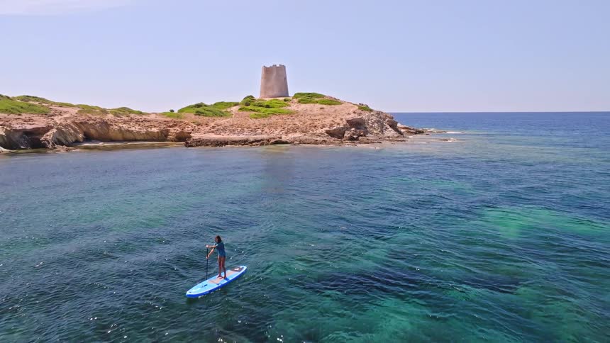 A woman on a paddleboard glides across the stunningly clear turquoise waters of a secluded bay, with an ancient coastal tower in the background