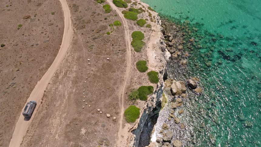 Aerial view of a silver van driving on a dirt road along a rugged cliffside with a beautiful turquoise ocean