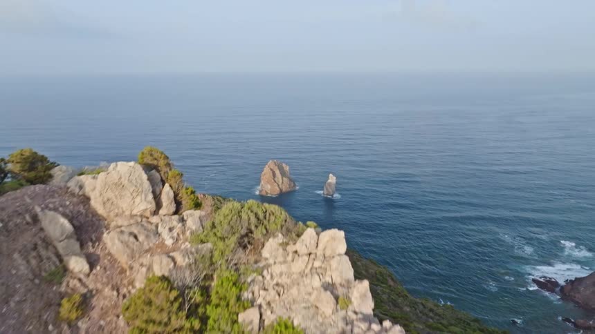 An aerial flyover reveals two stunning sea stacks rising from the ocean off a rugged, rocky coastline