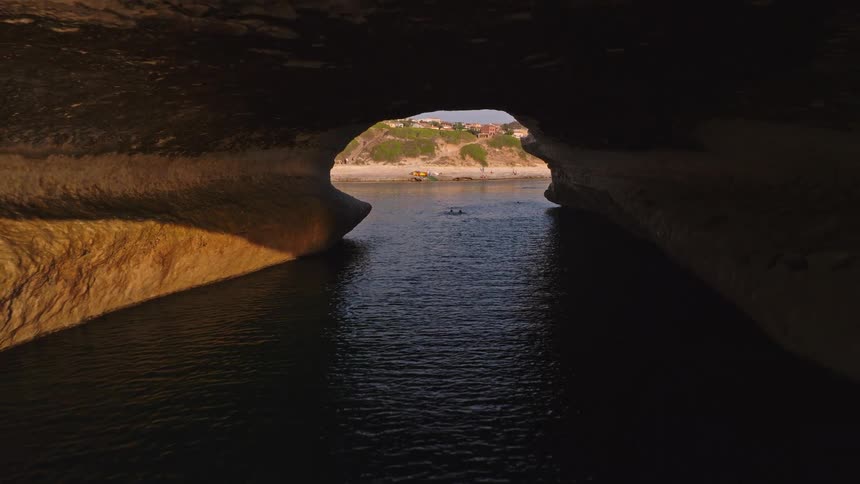 A stunning aerial shot pulls back out of a sea cave to reveal a magnificent natural rock arch over the ocean at golden hour