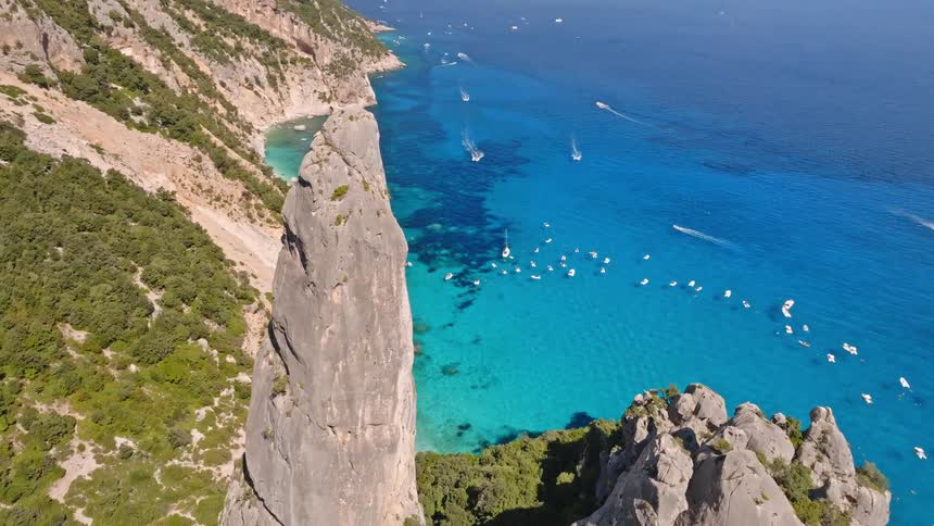 An aerial pullback reveals the iconic Aguglia di Goloritze sea stack standing sentinel over the brilliant turquoise bay of Sardinia, teeming with summer boats