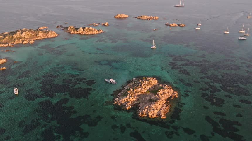 A serene aerial shot captures sailboats dotting the clear turquoise sea around a rocky archipelago at sunset