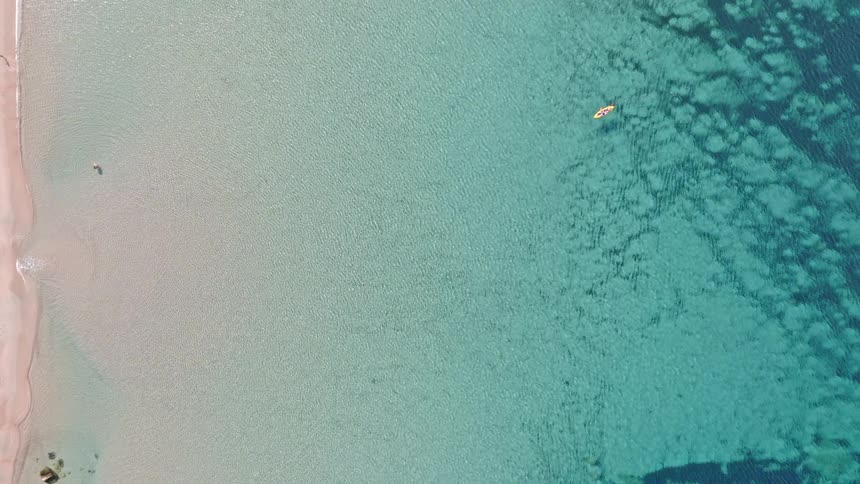 An aerial top-down view slowly zooms out to reveal a person kayaking in crystal clear turquoise waters next to a pristine sandy beach
