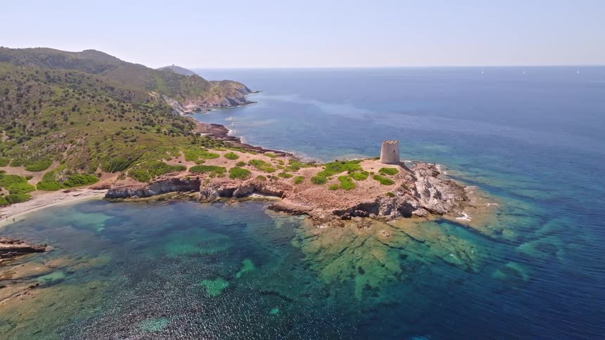 A beautiful aerial pullback shot reveals an ancient stone tower on a rocky peninsula along a stunning coastline with clear turquoise water