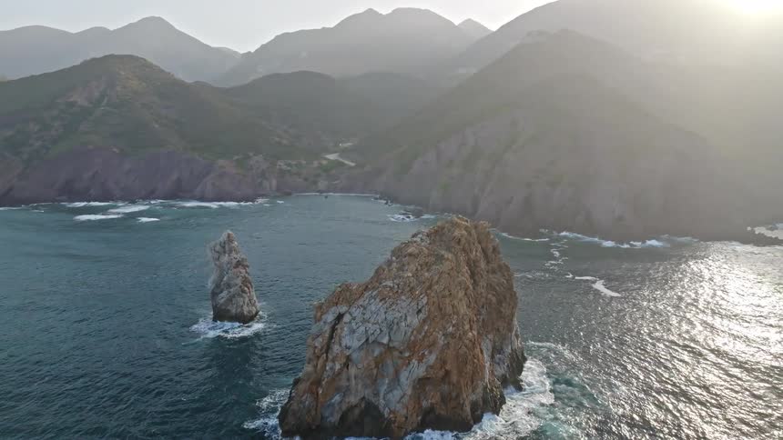 A majestic aerial view pulls back from a massive sea stack to reveal a stunning mountainous coastline at sunset