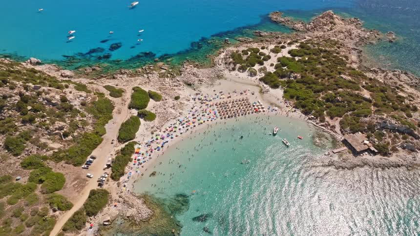 A breathtaking aerial view descends towards a bustling sandy cove filled with tourists enjoying the vibrant turquoise waters of a summer paradise