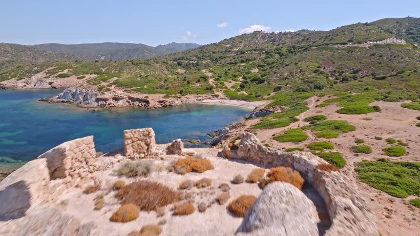 Aerial view flying over an ancient stone watchtower on a rugged cliff to reveal a stunning turquoise bay on a sunny day in Sardinia, Italy