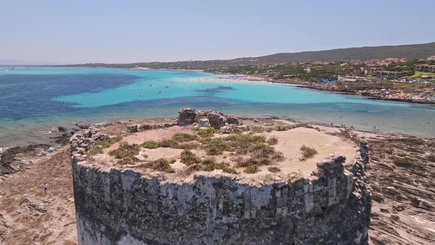 An aerial view slowly flies over a historic stone watchtower on a rocky coast, revealing a stunning turquoise bay and a busy beach on a sunny day