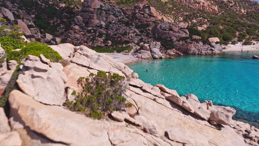A breathtaking aerial view moves over granite rocks to reveal the beautiful Cala Coticcio beach in Sardinia, with tourists swimming in the pristine turquoise sea