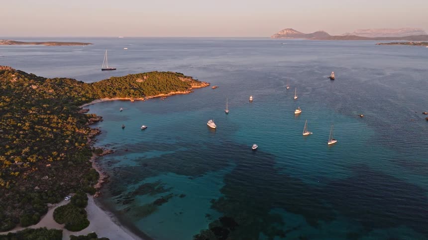 An idyllic aerial scene shows sailboats resting in a calm, clear turquoise bay surrounded by lush islands at sunset
