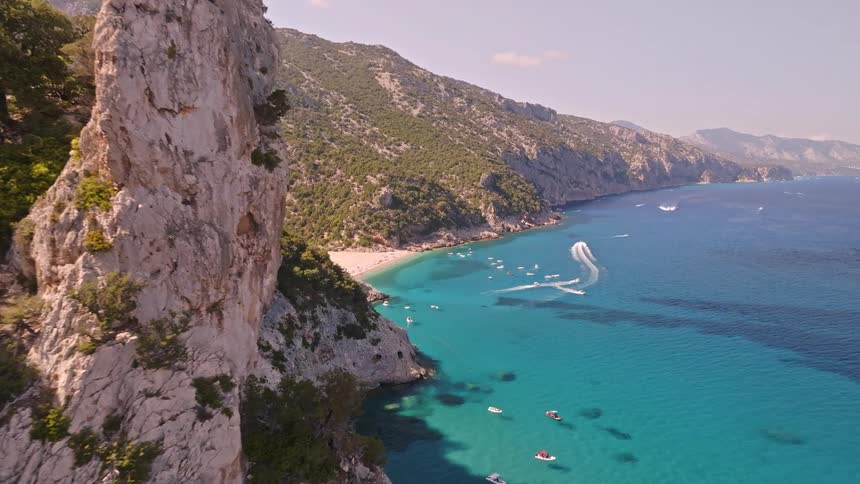 Flying between iconic sea stacks to reveal a breathtaking view of a secluded beach and turquoise ocean filled with boats on a sunny day