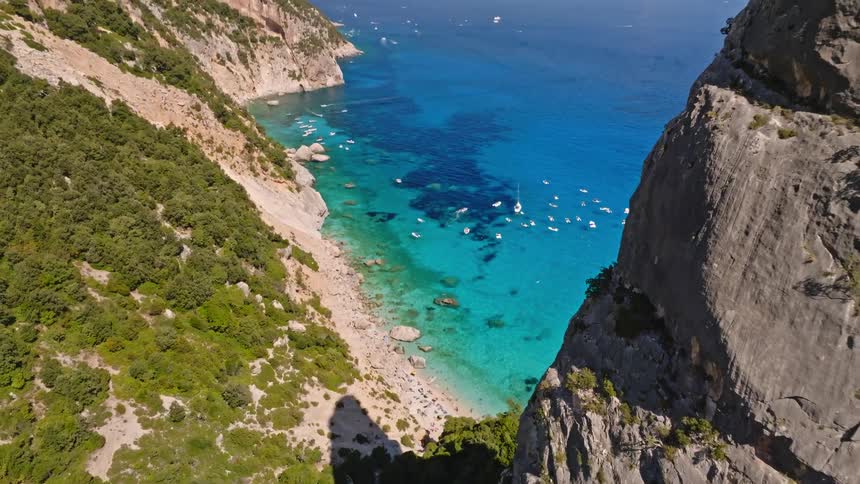 An aerial reveal shot of the breathtaking Cala Goloritzè, a secluded beach with turquoise waters and a towering rock pinnacle in Sardinia