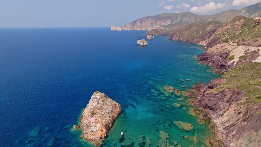 An aerial drone shot tilts up to reveal the stunning, rugged coastline and vibrant turquoise waters of a Mediterranean island on a sunny day