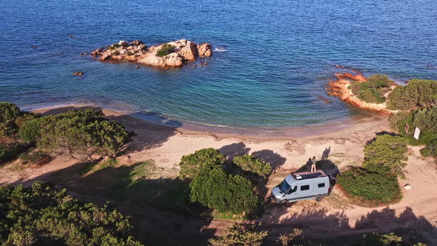 A stunning aerial view tilts up to reveal a couple standing atop their camper van, parked on a secluded beach overlooking the vast, beautiful ocean