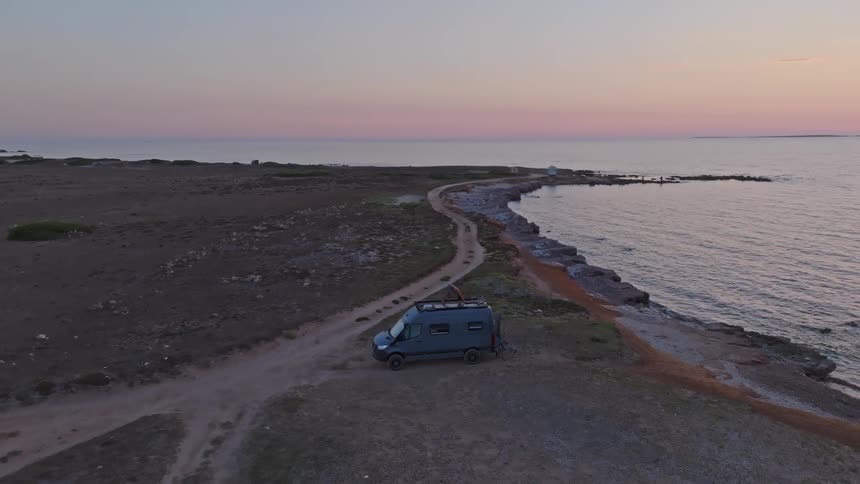 A person on top of a camper van enjoys the beautiful ocean sunset from a remote, rocky coastline