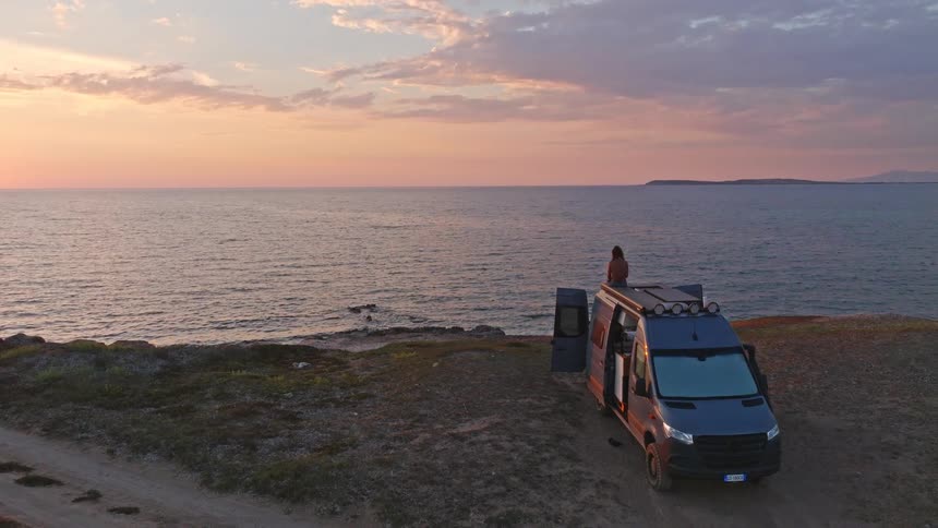 A lone traveler sits on top of their camper van parked on a coastal cliff, peacefully watching a beautiful sunrise over the calm ocean
