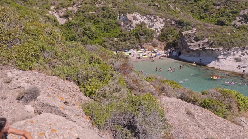 A young woman sits peacefully on a rocky cliff, admiring the breathtaking view of a secluded cove where people swim and relax on the beach